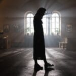 A woman stands silhouetted in an atmospheric orthodox church, light streaming through windows.