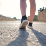 A person walks on a sunny rural road wearing sneakers, casting a long shadow.