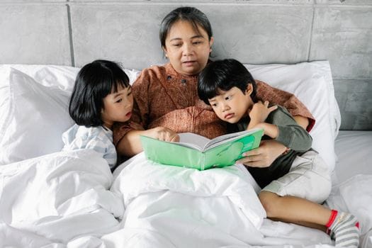 From above of Asian grandmother lying with little daughter and son during bed time and reading book