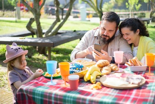 Happy family having a picnic in a sunny park, enjoying snacks and using a smartphone together.